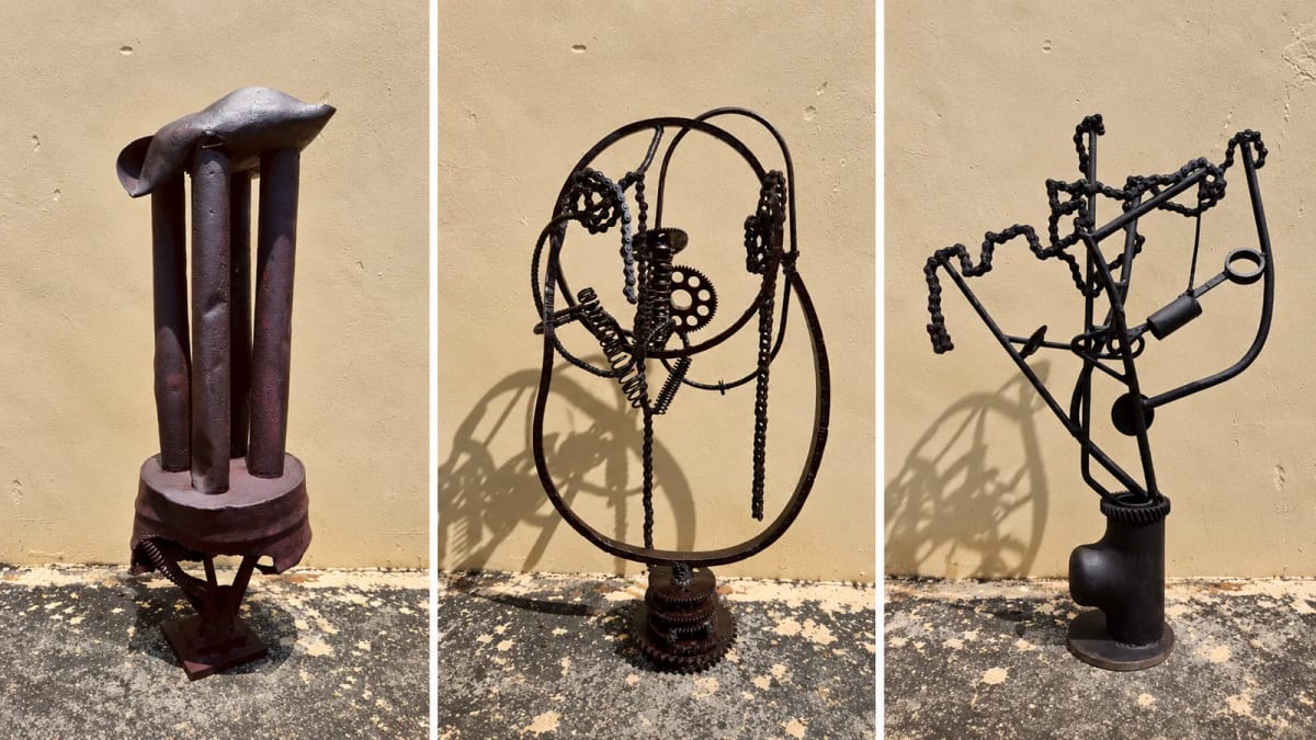 Three metal sculptures assembled from rods, gears, springs and bicycle chains stand against a yellow wall.