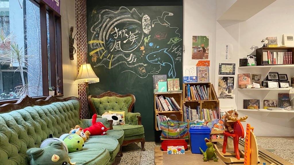 Stuffed animals occupy a green sofa beside low picture-book shelves, a rocking horse, a standing lamp and a hand-drawn blackboard.