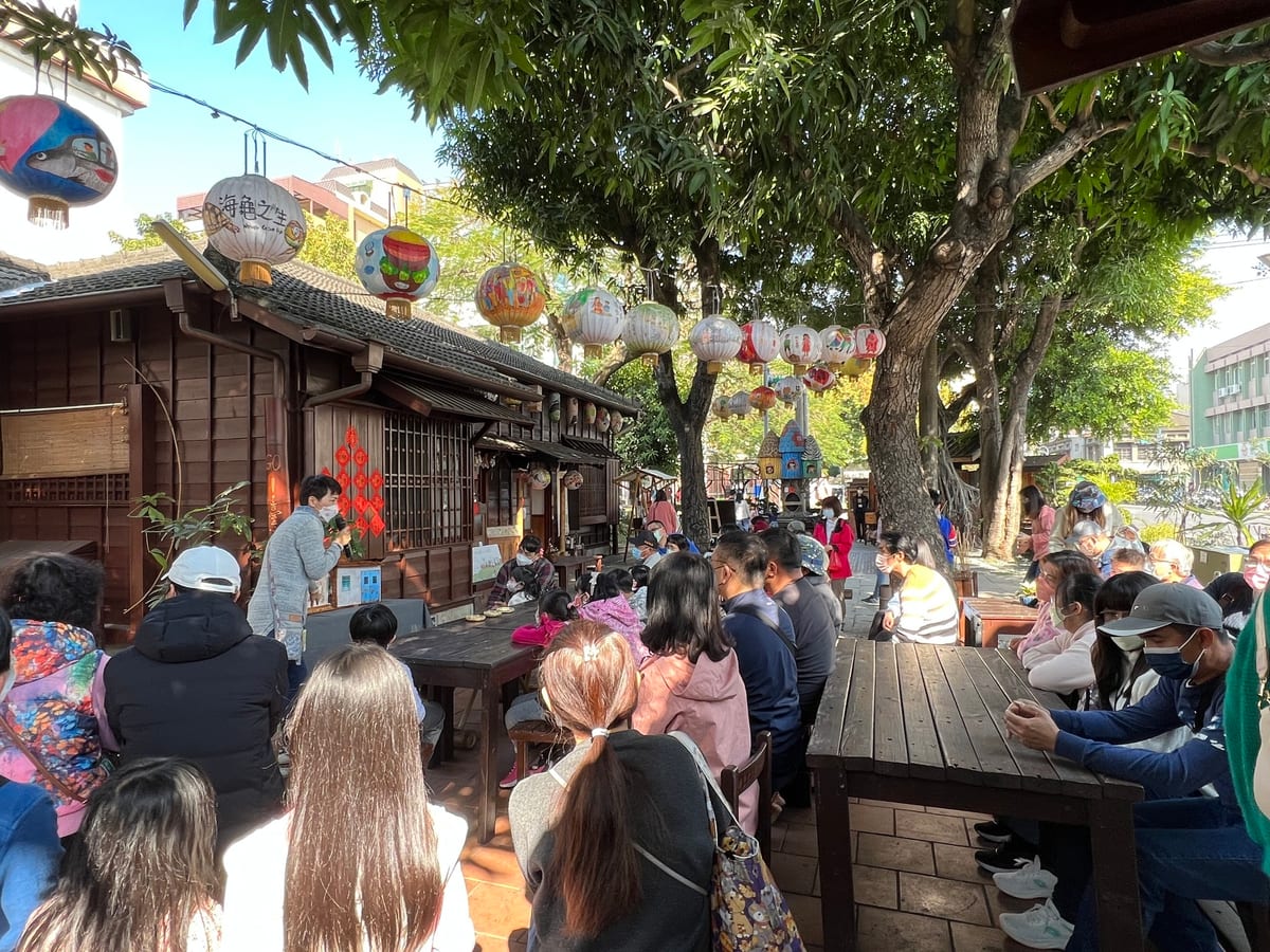 An audience sits at wooden tables beneath a large tree and painted lanterns beside Yunlin Storyhouse's dark wooden walls and tiled roof.