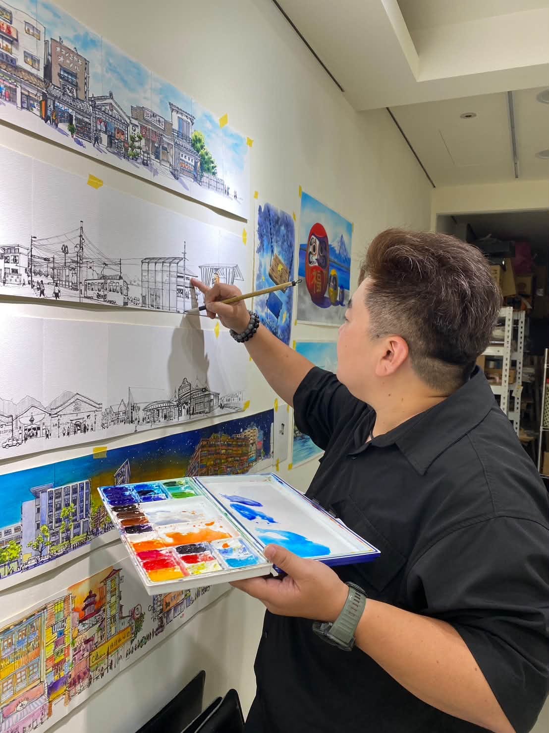 An artist with a palette paints beside long city sketches on the gallery wall.